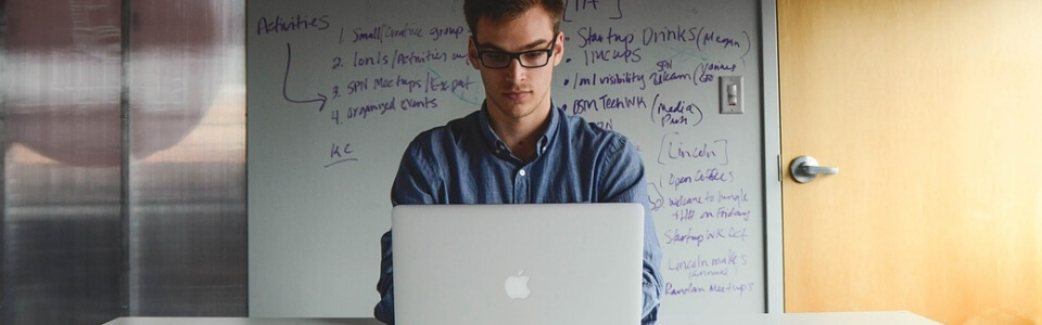 Ein Projektmanager im Bereich Sportsponsoring arbeitet an einem Laptop im Hintergrund ist ein beschriftetes Whiteboard zu erkennen