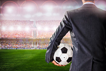 Mann im Anzug steht im Stadion und hält Fußball unter dem Arm.